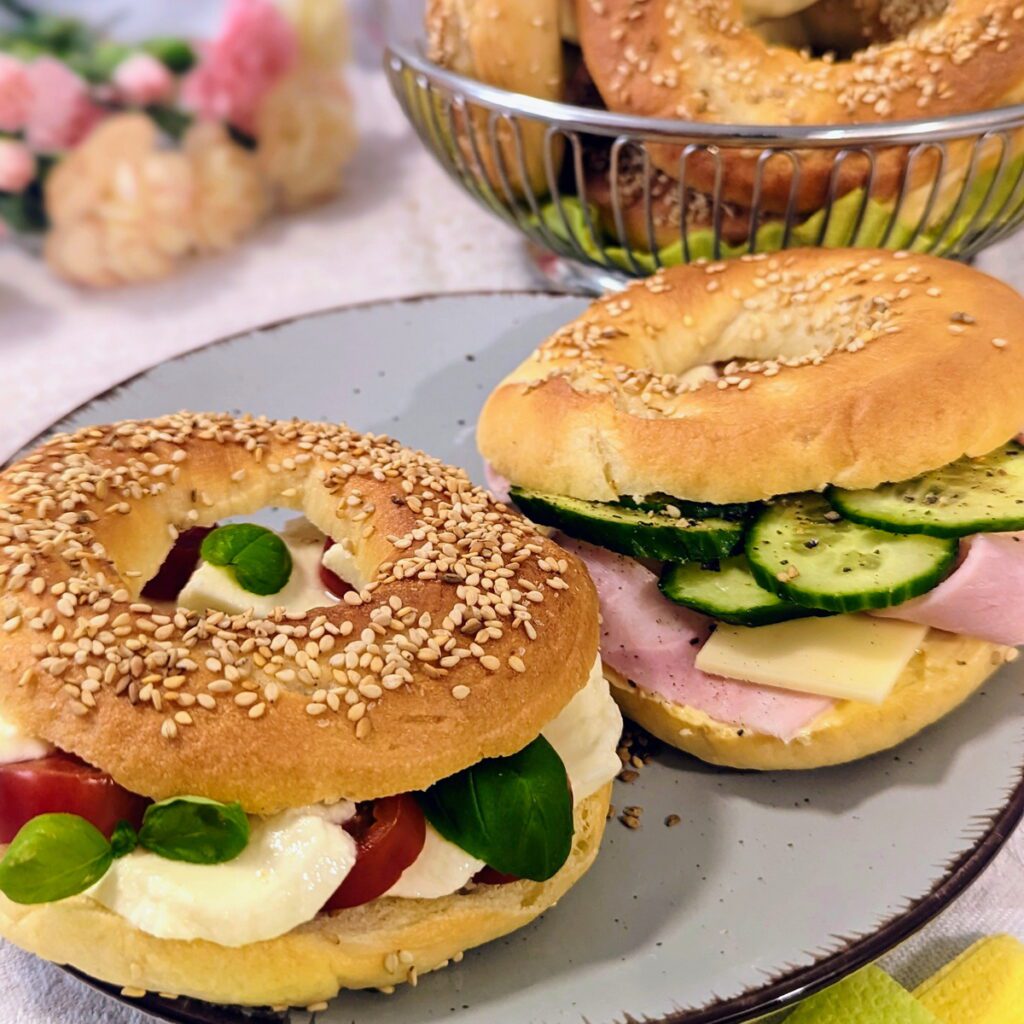 Zwei belegte Bagels, mit Tomate-Mozzarella und Schinken-Käse und Gurken. Im Hintergrund steht ein Korb mit frischen unbelegten Bagels