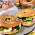 Zwei belegte Bagels, mit Tomate-Mozzarella und Schinken-Käse und Gurken. Im Hintergrund steht ein Korb mit frischen unbelegten Bagels