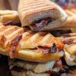 Köstliche Double-Cheese-Bacon Panini, perfekt goldbraun gegrillt und mit saftigen Tomatenscheiben gefüllt. Die Panini lieben auf einem rustikalen Holzbrett