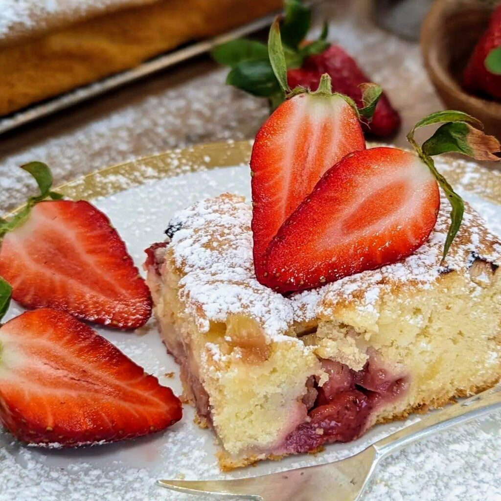 Ein saftiger Erdbeer-Rhabarber-Kuchen vom Blech, dekoriert mit frisch geschnittenen Erdbeeren, bestäubt mit Puderzucker.