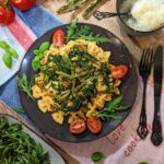 Ein Teller mit Farfalle, frischem grünem Spargel, saftigen Tomaten und cremigem Mozzarella, garniert mit frischem Basilikum und serviert auf einem rustikalen Holztisch neben einer Schüssel Rucolasalat.