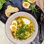 Ein Teller mit dampfender Schlesischer Sauerampfer-Suppe, garniert mit gekochten Eiern und frisch geschnittenem Sauerampfer. Dazu serviert wird knuspriges Brot.