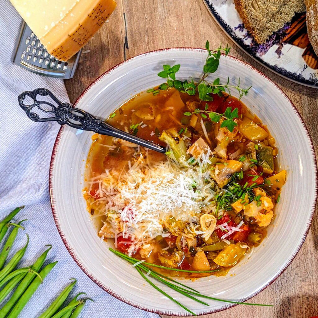 Ein dampfender Teller Gemüsesuppe mit italienischen Kräutern, garniert mit frischem Parmesan, Schnittlauch und einem Zweig Oregano und serviert mit einer Scheibe rustikalem Brot