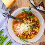 Ein dampfender Teller Gemüsesuppe mit italienischen Kräutern, garniert mit frischem Parmesan, Schnittlauch und einem Zweig Oregano und serviert mit einer Scheibe rustikalem Brot