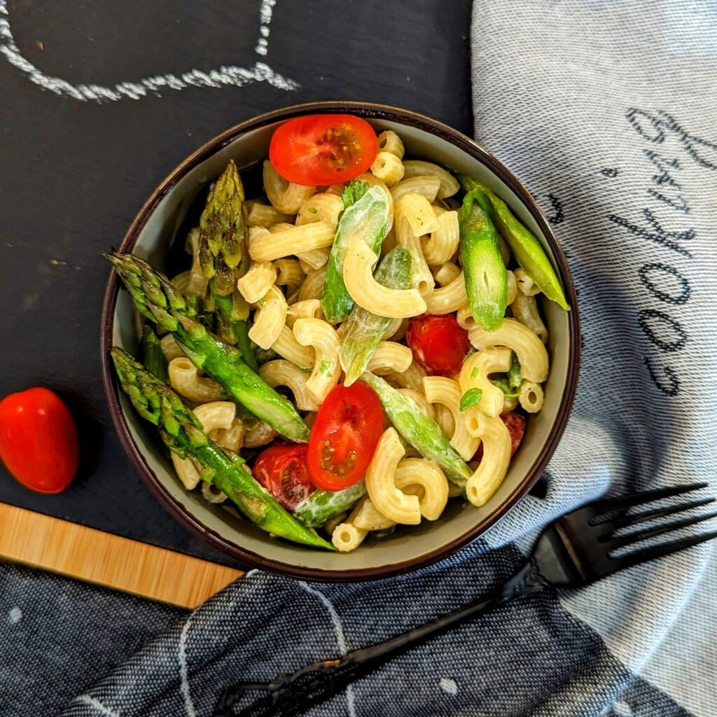 Ein dekorativer Teller mit Nudelsalat, grünem Spargel, Tomaten und frischen Kräutern, serviert auf einer beschrifteten Schieferplatte