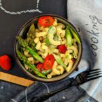 Ein dekorativer Teller mit Nudelsalat, grünem Spargel, Tomaten und frischen Kräutern, serviert auf einer beschrifteten Schieferplatte