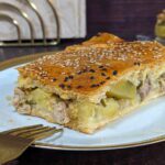 Eine goldbraune, knusprige Pastete, liebevoll mit einem Teigmuster verziert und mit einem glänzenden Eigelbüberzug., bestreut mit Sesam und Schwarzkümmel
