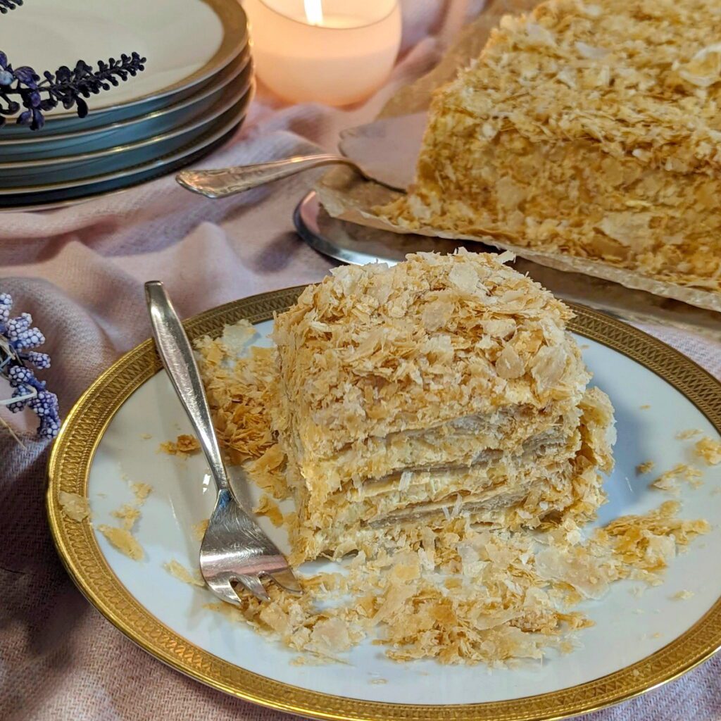 Ein üppiges Stück Kuchen aus schichtweise angeordnetem knusprigem Blätterteig und cremiger Butter-Kondensmilch-Creme, verziert mit zarten Blätterteig-Bröseln, der auf einem Kuchenteller serviert wird.