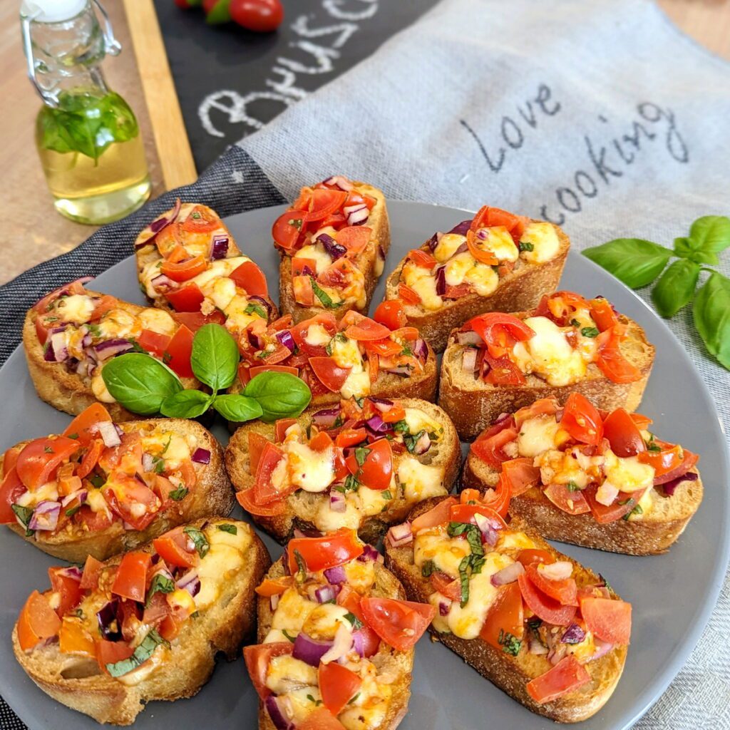 Dekorative Tomaten-Mozzarella Bruschetta auf einem Teller, garniert mit frischen Basilikumblättern und einer Prise schwarzem Pfeffer. Im Hintergrund ein Fläschchen mit Basilikumöl